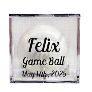 May include: A clear acrylic box encases a white game ball. The ball features the name "Felix", the words "Game Ball", and the date "May 17th, 2025" in black script. The box is displayed against a white background.