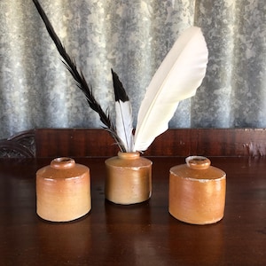 May include: Three antique ceramic inkwells in shades of brown, with a rustic, aged appearance. One inkwell holds a collection of feathers, including a large white feather and several black ones. The inkwells are arranged on a dark wooden surface.