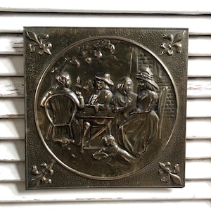 May include: A square, bronze-coloured metal wall plaque featuring a detailed relief scene of figures around a table. The scene is framed by a circular border and fleur-de-lis corner accents. The plaque is mounted on a white slatted surface.