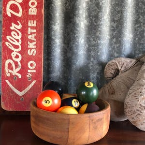 May include: A wooden bowl containing various billiard balls, including orange, black, green, and yellow, each numbered. A red sign with white text reading "Roller Derby IO Skate Board" is visible on the left.