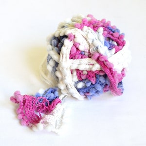 Puede incluir: Una bola de hilo con pompones blancos, rosas y azules. El hilo es una mezcla de blanco, rosa y azul.