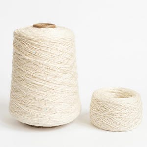Op de afbeelding: Een grote, hoge, conische klos garen naast een kleinere, ronde, platte bol van hetzelfde gebroken witte garen. Beide items hebben een getextureerd, vezelig uiterlijk met een consistente lichte crèmekleur door de vezels heen.