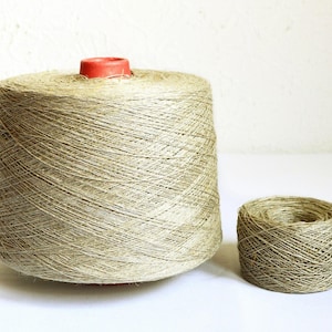 May include: A large, conical spool of natural, light brown fiber yarn with a red plastic core at the top, positioned next to a smaller, cylindrical spool of the same textured, earthy-toned yarn, both featuring a visible, cross-wound thread pattern.