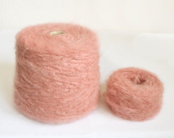 Puede incluir: Dos carretes de hilo suave de color rosa. El carrete más grande está lleno, mientras que el carrete más pequeño está parcialmente desenrollado.