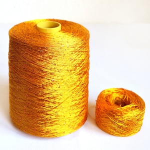 Puede incluir: Una bobina de hilo dorado con un acabado brillante. El hilo está enrollado alrededor de una bobina de plástico amarilla.