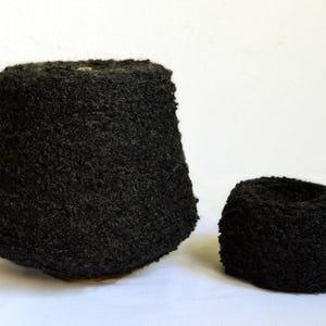 Pode incluir: Dois cones de fio preto texturizado são exibidos em um fundo branco. O cone maior é de cor carvão escuro, e o cone menor é de um tom semelhante. O fio parece espesso e macio, adequado para projetos de tricô ou crochê.