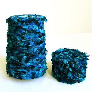 Könnte beinhalten: Zwei Garnspulen in Blau- und Schwarztönen. Die größere Spule ist kegelförmig, während die kleinere zylindrisch ist. Das Garn hat ein strukturiertes, blattartiges Aussehen und erzeugt einen visuell interessanten Kontrast.