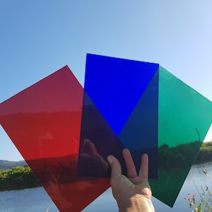 Op de afbeelding: Een hand houdt drie transparante gekleurde vellen vast tegen een blauwe lucht en water achtergrond. De vellen zijn rood, blauw en groen. Het blauwe vel heeft een driehoekige vorm. De foto is buiten gemaakt.