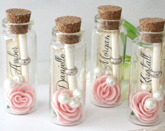 May include: Set of small glass bottles with cork stoppers, each containing a rolled piece of paper, a pink fabric rose, and white pearl beads. The bottles are arranged on a white surface with a card that reads "Whitney, I can't say I do without you. Will you be my Bridesmaid? Love, Tiffany."