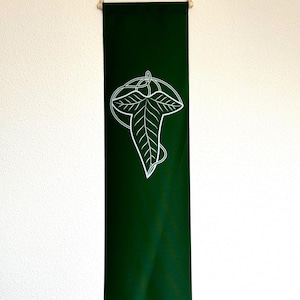 Könnte beinhalten: Ein grünes Stoffbanner mit einem weißen Blattdesign. Das Blatt ist stilisiert und hat oben einen Knoten. Das Banner hängt an einer Holzstange.