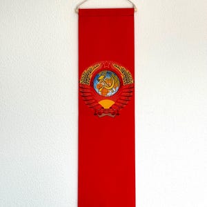 Op de afbeelding: Een rode vlag met een gouden embleem met een hamer en sikkel, een wereldbol en tarwehalmen. Het embleem is gecentreerd op de vlag, die aan een witte muur hangt.