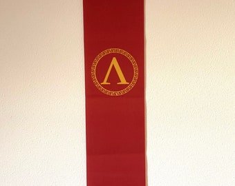 May include: A red fabric banner with a gold Greek letter 'A' inside a circle with a gold Greek key pattern.