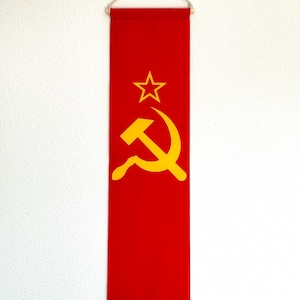 May include: A red fabric banner with a yellow hammer and sickle and a yellow star. The banner is hanging from a white string.