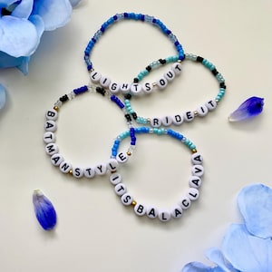 May include: Four circular beaded bracelets featuring blue, light blue, and black seed beads with white letter beads. The bracelets display the phrases LIGHTS OUT, RIDE IT, BATMAN STYLE, and ITS BALACLAVA in black text on the white beads.