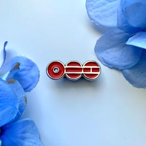 May include: A horizontal metal pin featuring three circular segments with a bright red enamel fill and silver-toned edges. The first circle contains a small central hole, while the second and third circles feature horizontal silver bars.