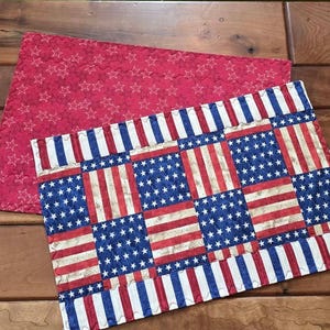 Op de afbeelding: Twee patriottische placemats. Eén is rood met witte sterren. De andere heeft een patchwork-ontwerp van de Amerikaanse vlag met rode en witte strepen en blauwe velden met witte sterren. De placemats zijn gewatteerd.