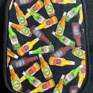 May include: Black fabric pot holder with a repeating pattern of various beer bottles. The bottles are labeled with different beer types, including "Lager", "Pilsner", "Ale", and "Stout".