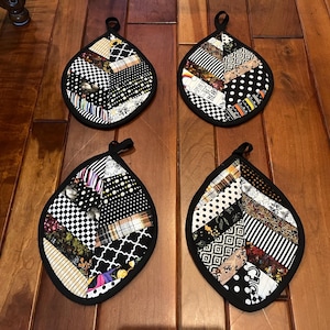 May include: Four black and white leaf-shaped pot holders with a patchwork design. The pot holders are made of different fabrics with black, white, brown, and gold patterns.
