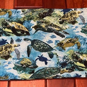 Op de afbeelding: Een blauwe en groene stof met een herhalend patroon van zeeschildpadden die in een koraalrif zwemmen. De stof heeft een verscheidenheid aan zeeschildpadden in verschillende maten en posities, sommige met het gezicht naar de kijker en andere die wegzwemmen. De stof is perfect voor quilten, knutselen en interieurdecoratie.
