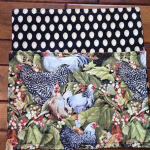 May include: A set of two fabric placemats. The top placemat is black with white polka dots. The bottom placemat features a colorful pattern of chickens and green leaves.
