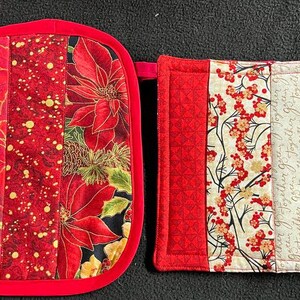 Puede incluir: Dos agarraderas de olla acolchadas de color rojo y blanco. Una agarradera presenta un patrón de poinsettia roja y la otra tiene un patrón floral rojo y blanco con el texto "Together" repetido.