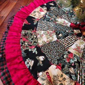 May include: A Christmas tree skirt with a patchwork design. The skirt has a red ruffled border and a plaid outer edge. The patchwork includes bird, floral, and striped patterns, in red, black, and cream colours.