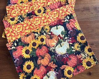 May include: A set of rectangular fabric placemats featuring a repeating pattern of orange and white pumpkins, yellow sunflowers, and autumn leaves on a black background, with a contrasting orange and yellow textured fabric border.