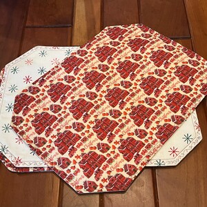 Puede incluir: Dos manteles individuales octogonales con fondo blanco y copos de nieve bordados en rojo y verde. Un mantel individual tiene un patrón rojo y blanco de casas de jengibre y la palabra "Navidad" repetida en todo.