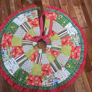 May include: A colorful patchwork Christmas tree skirt with a red, green, and white color scheme. The skirt is made of fabric scraps and has a ruffled edge. It features a variety of patterns, including polka dots, stripes, and snowflakes.