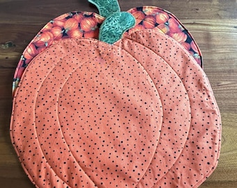 May include: A quilted fabric pumpkin-shaped mat featuring a light orange surface with small black dots and a green stem. A second layer is visible underneath, showcasing a dark fabric with a repeating orange pumpkin pattern and green stems.