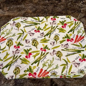 May include: White octagonal placemat with a quilted design featuring a variety of vegetables, including asparagus, artichokes, radishes, carrots, and peas. The placemat has a white background with green and brown accents. The text "Sugar Snap Peas", "Asparagus", "Carrots", "Radish", "Artichoke", and "Carrots" is printed on the placemat.