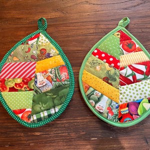 Puede incluir: Dos agarraderas acolchadas en forma de hoja con ribete verde y presillas para colgar. Las agarraderas presentan un diseño de patchwork con varias telas, incluyendo rayas rojas y blancas, lunares e impresiones de verduras.