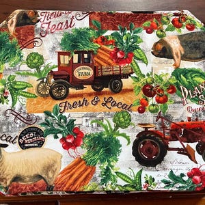May include: A white fabric placemat with a red truck carrying vegetables, a pig, a sheep, and a red tractor. The placemat has a farmhouse theme with the text "Fresh & Local" and "Plant Seed Company".
