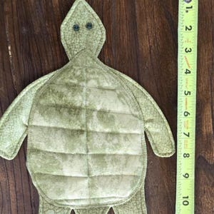May include: A green fabric turtle hot pad with a textured surface and black button eyes. The hot pad is approximately 7 inches long.