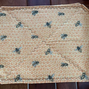May include: A rectangular placemat featuring a honeycomb design and bee motifs. The colour scheme is yellow and orange, with black and white bees. The edges are scalloped and stitched with decorative thread.