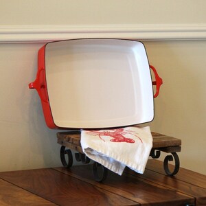 May include: A rectangular enameled cast iron baking dish with a bright red exterior and a smooth white interior. The dish features two integrated side handles and is positioned upright, accompanied by a white cloth featuring a red lobster graphic.