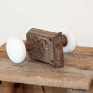 May include: An antique door lock mechanism featuring a rectangular, rusted metal plate with intricate embossed scrollwork patterns. Two smooth, round, white porcelain knobs are attached to the central spindle on either side of the metal housing.