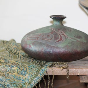 May include: A handcrafted, oval-shaped ceramic vase with a dark green and burgundy finish. The vase features a carved design of a bird. The vase is displayed on a wooden surface next to a patterned scarf.
