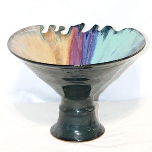 May include: A ceramic pedestal bowl with a flared, dark teal base and a wide, wavy rim. The interior rim features a colorful, vertical stripe design in shades of yellow, red, purple, blue, and teal. The bowl has a glossy finish.