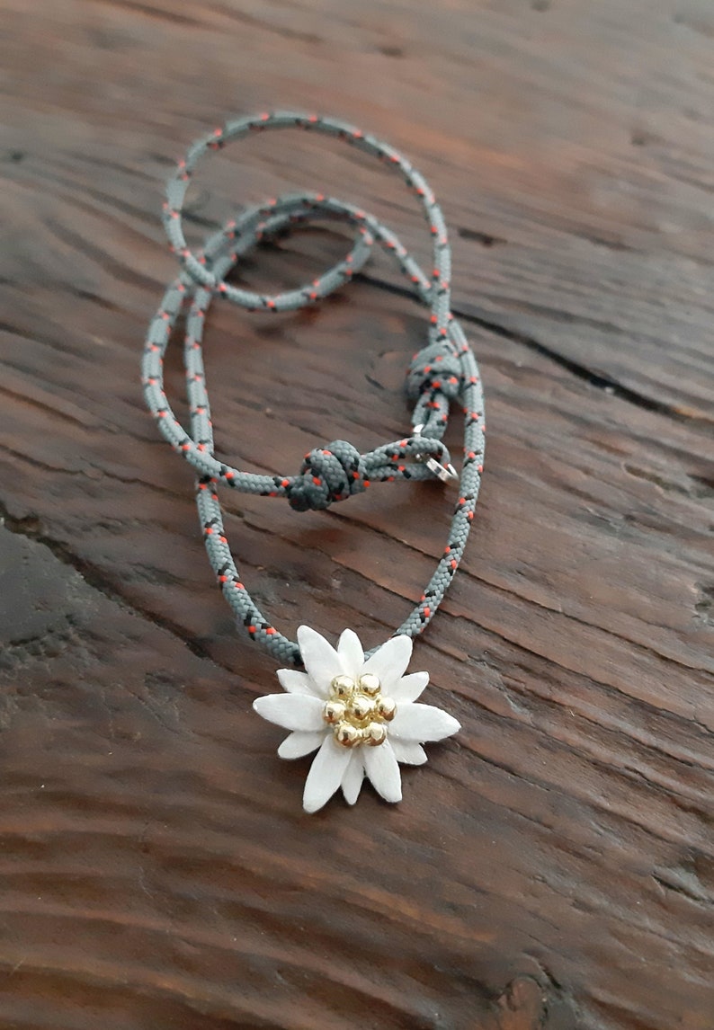 May include: A gray and orange rope necklace with a white edelweiss flower pendant. The flower has a gold center with small gold beads.