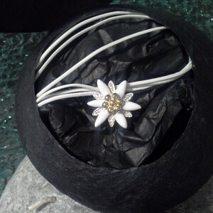 May include: A white edelweiss flower charm with gold accents is nestled in a black velvet box with white satin ribbon.