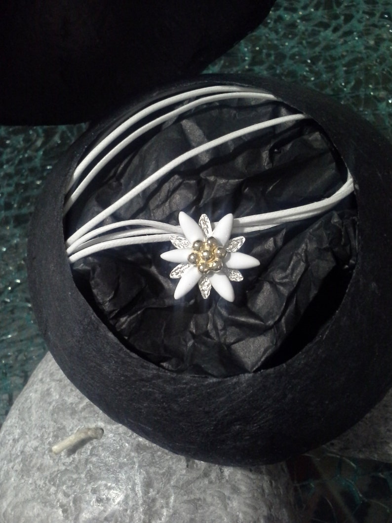 May include: A white edelweiss flower charm with gold accents is nestled in a black velvet box with white satin ribbon.