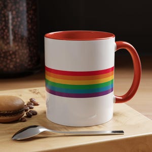 May include: A white ceramic mug with a red rim and handle. The mug has a rainbow stripe design on the front.