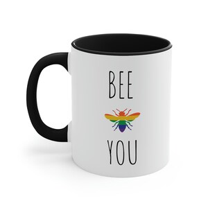 May include: White ceramic coffee mug with a black rim and handle. The mug has the text "BEE YOU" printed on it in black with a rainbow bee illustration in the center.