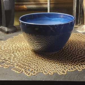 May include: A deep blue ceramic bowl with a textured surface sits on a gold-colored placemat. The bowl is round and has a wide rim.
