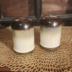 May include: A pair of ceramic salt and pepper shakers. The shakers are cylindrical with a glossy, dark brown top that fades into a creamy white base. The shakers sit on a gold-colored, patterned surface.