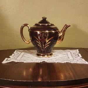 May include: A dark brown teapot with gold accents and a floral design. The teapot has a round body and a spout on the side. It is sitting on a white lace doily.