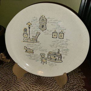 May include: A white oval ceramic plate with a black and yellow design of a brick wall with a rocking chair, a lamp, a piano, and other home decor items.