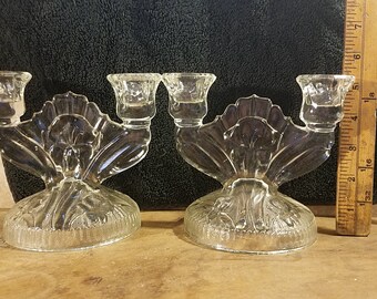 May include: Two clear glass candle holders with a double-candle design. Each holder features a fan-shaped back and a textured base. A wooden ruler is positioned next to the candle holders.