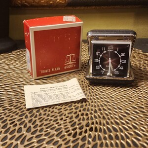 May include: A vintage Equity travel alarm clock with a black face, white numerals, and a red second hand. The clock is in a black case, next to its original red and white box. The box reads "TRAVEL ALARM EQUITY".